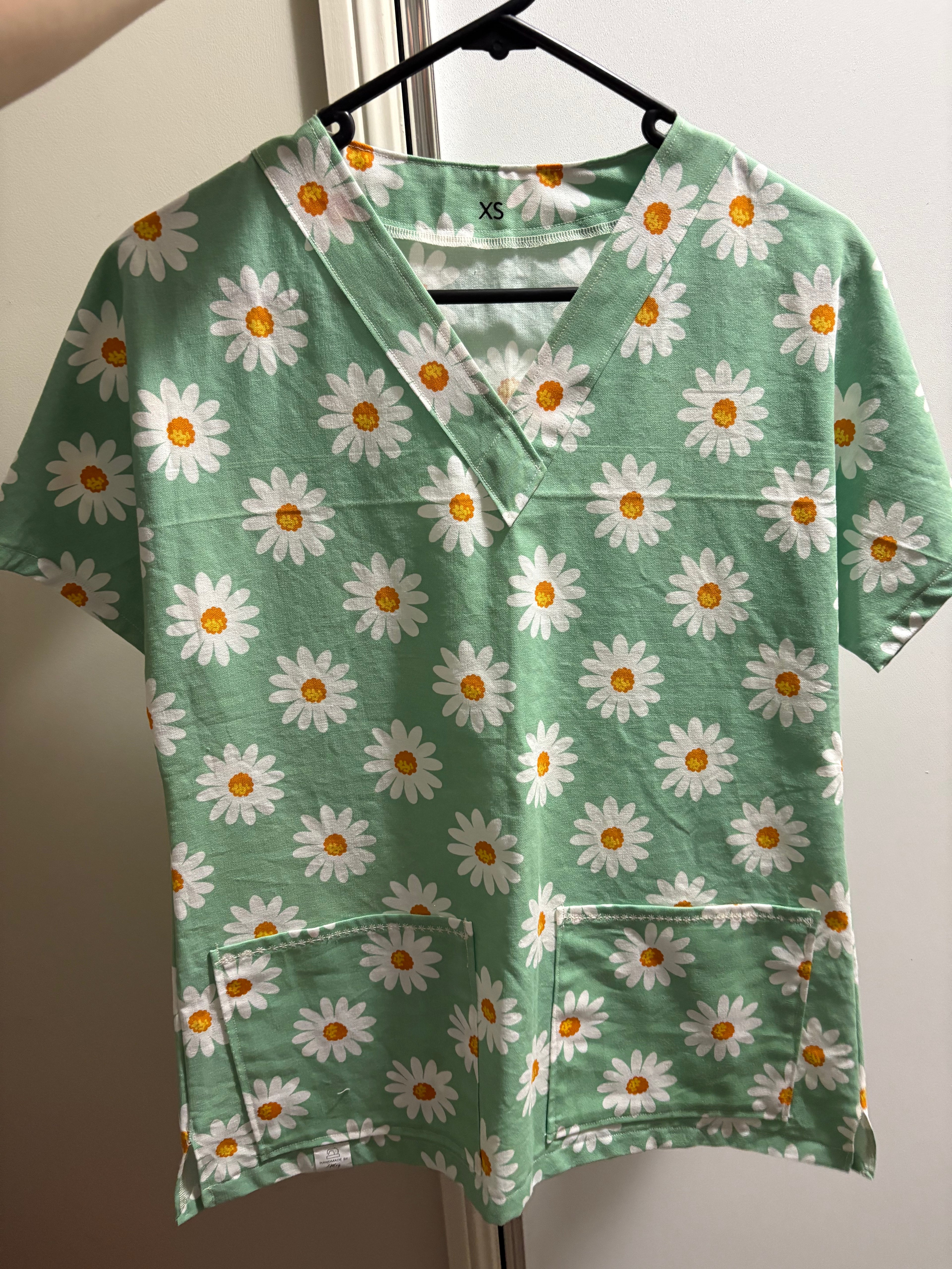 Green scrub top with white daisy pattern on a hanger against a neutral background